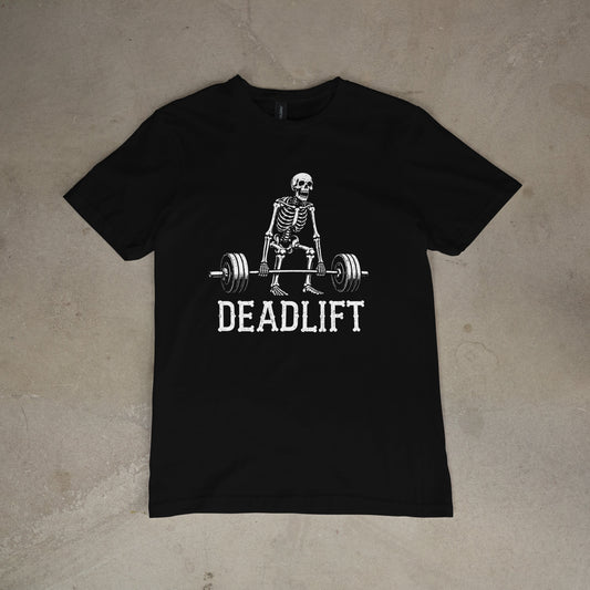 A black short-sleeve crewneck t-shirt laid flat on a textured concrete surface, featuring a white graphic of a skeleton lifting a barbell with the word 'DEADLIFT' printed underneath.