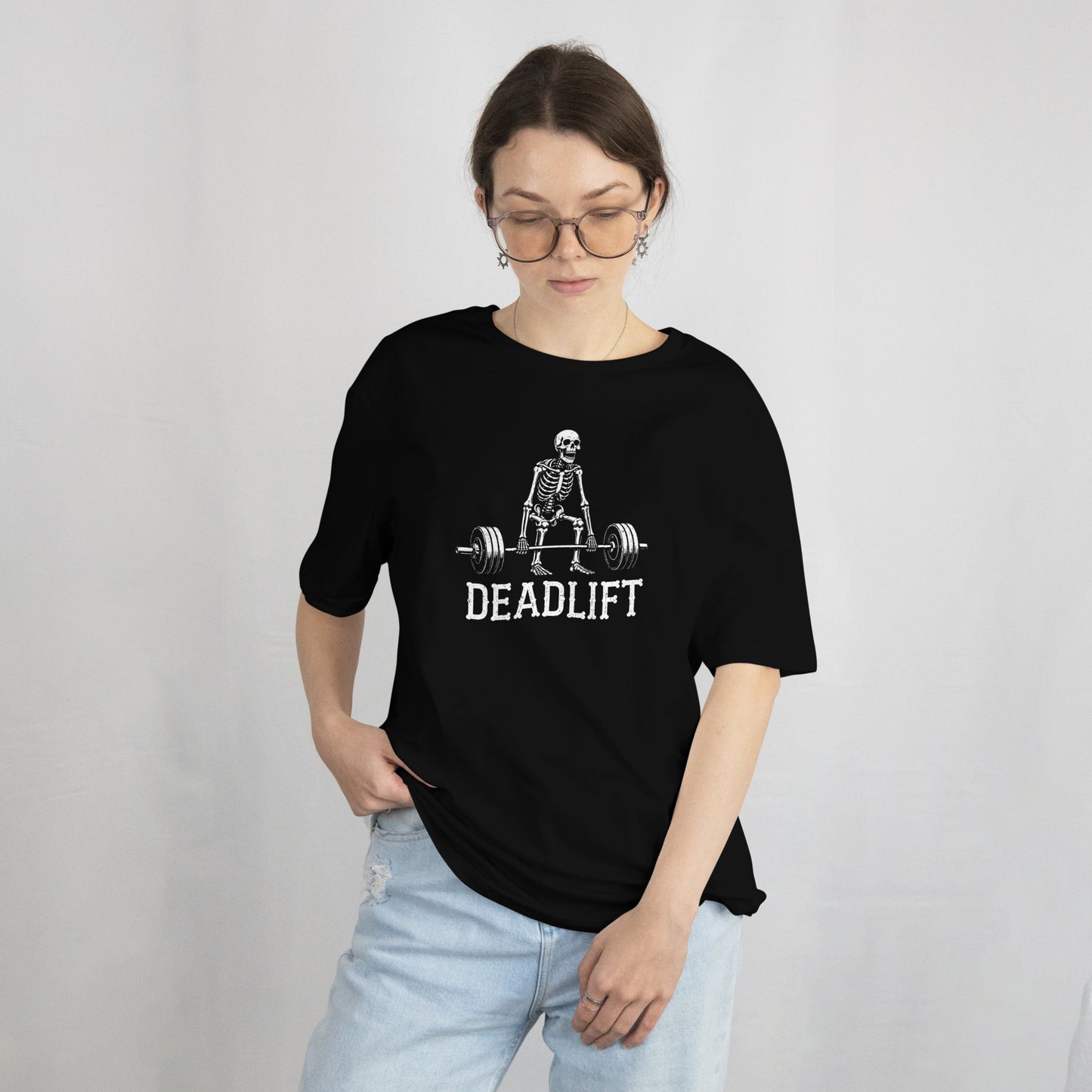 A woman wearing a black oversized t-shirt with a white graphic of a skeleton lifting a barbell and the word 'DEADLIFT' printed below it, paired with light blue jeans, set against a plain light gray background.