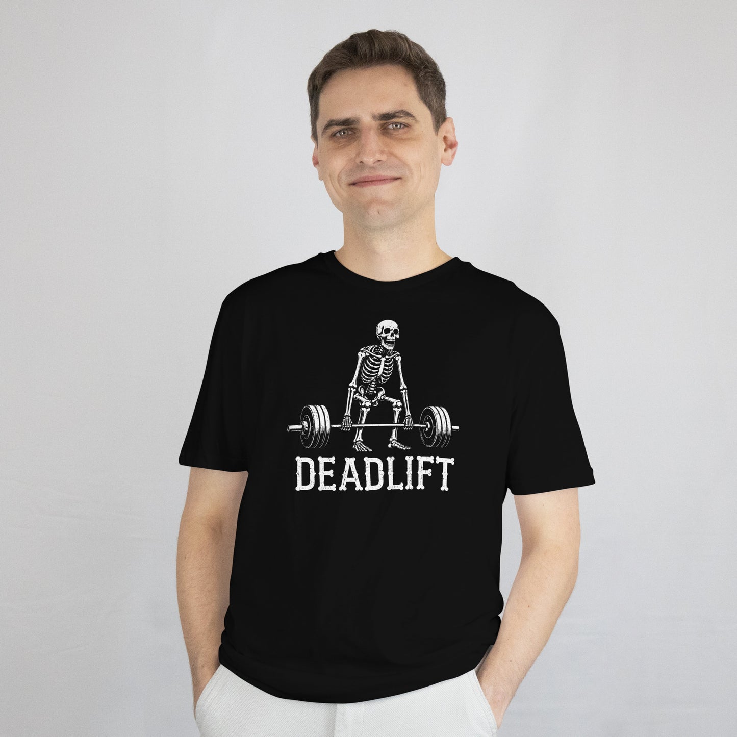 A man wearing a black crewneck t-shirt with a white graphic of a skeleton lifting a barbell and the word 'DEADLIFT' printed below it, paired with white pants, against a light gray studio background.